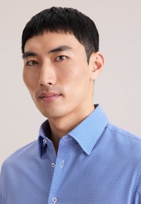 Niebieska koszula zapinana na guziki z geometrycznym wzorem, ze sztywnym kołnierzykiem i gładką fakturą. Lekka tkanina, dopasowany krój.