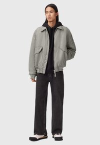 Chaqueta bomber gris con grandes bolsillos frontales, usada sobre un hoodie negro, combinada con jeans de pierna ancha negros y zapatillas blancas con suelas negras.