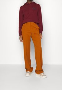 Sudadera con capucha color burdeos con cordones y logo, combinada con pantalones naranjas de cintura alta y pierna ancha, y zapatillas de deporte beige.