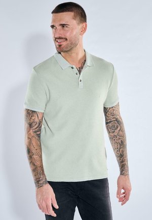 Mann mit Ärmeltätowierungen trägt ein hellgrünes, strukturiertes Poloshirt und schwarze Hose, schaut nach links und steht vor einem schlichten hellen Hintergrund.