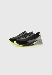 Zwarte en grijze sportschoenen met een mix van materialen, opvallende veters en felle gele accenten op de zool en hiel.