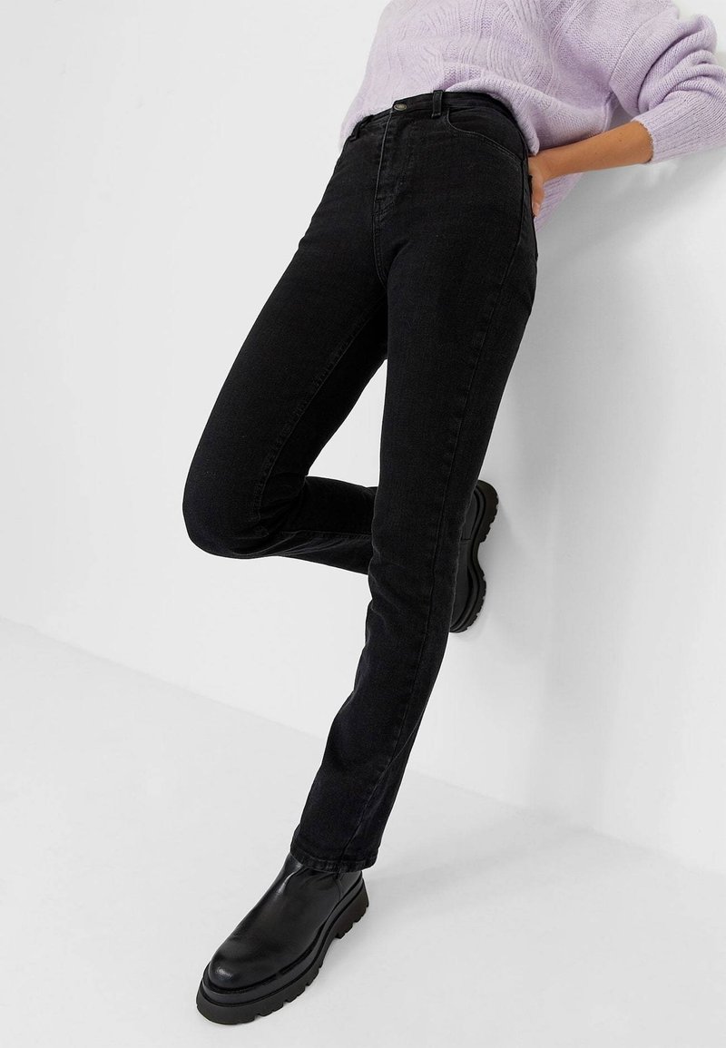 Personne portant un jean noir slim, des bottes noires épaisses et un pull en tricot lavande, appuyée contre un mur blanc.