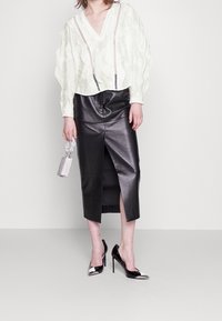 Blusa bianca strutturata con scollatura a V profonda, gonna a tubino nera in ecopelle con spacco, tacchi a punta neri e argento, e una borsa metallica.