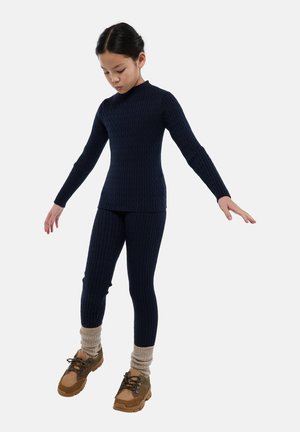Jeune personne en équilibre sur une jambe, portant un pull en maille bleu marine et des leggings, des guêtres beige, et des chaussures de randonnée marron.