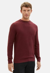 Burgunderfarbener Strickpullover mit Rundhalsausschnitt, langen Ärmeln, strukturiertem Stoff und gerippten Bündchen. Getragen mit schwarzen Hosen, die eine taillierte Passform zeigen.