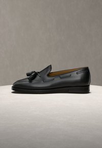 Fekete bőrs mokaszin bojttal, alacsony sarokkal és varrott részletekkel, világos felületen, bézs háttér előtt elhelyezve.