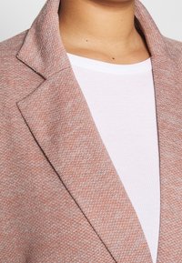 Vaaleanpunainen teksturoitu bleiseri, jossa on lovettu kaulus ja tyköistuva malli, kerroksittain valkoisen ribbipaidan päällä. Lähikuva korostaa materiaalin yksityiskohtia.