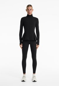 Zwarte sportieve jas met hoge kraag en peplumdesign, gecombineerd met zwarte legging en witte sneakers. Glad, rekbaar materiaal.