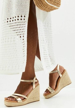 Ciemnoskóre nogi w białej szydełkowej spódnicy i złotych koturnach z paskami wokół kostek, trzymające pasujący słomkowy kapelusz.