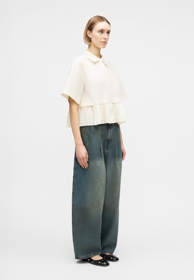 Γυναίκα που φοράει λευκό πλισέ πουκάμισο με κοντά μανίκια, φαρδιά ξεβαμμένα μπλε τζιν και μαύρα μπαλαρίνια με ασημένιες τρουκς, στέκεται προφίλ.