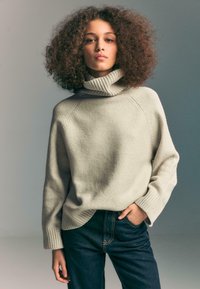 Donna con capelli ricci, che indossa un maglione beige a collo alto e jeans blu scuro, in posa con una mano in tasca su sfondo grigio.
