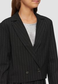 Blazer noir à rayures avec des accents argentés, design croisé, revers crantés et tissu lisse, porté sur un haut argenté.