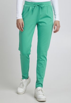 Personne portant un pantalon de jogging vert ajusté avec des poches avant et des baskets blanches, debout devant un fond uni.