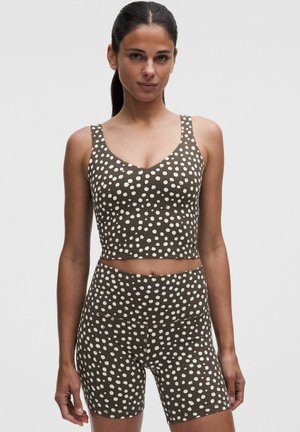 Kvinde iført matchende brun og hvid prikket ærmeløs crop top og højtaljede biker shorts mod en ensfarvet baggrund.