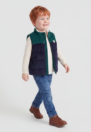 Junges rothaariges Kind lächelt und geht, trägt eine grüne und marineblaue Weste, ein cremefarbenes Langarmshirt, blaue Jeans und braune Stiefel.