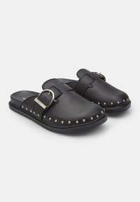 Musta nahkaiset slip-on-kengät, joissa on leveä hihna, jossa on hopeinen solki ja hopeiset niitit pohjan reunassa. Tasapohjainen muotoilu.
