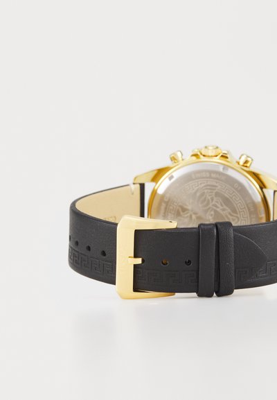 Zelta tonis hronogrāfa pulkstenis ar melnu ādas siksniņu, kurai ir iegravēts grieķu atslēgas raksts un zelta sprādze, atklājot rakstainu aizmuguri.