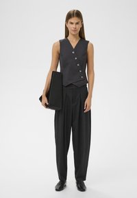 Šedý bezrukávový sveter s piatimi gombíkmi, kombinovaný s tmavými voľnými nohavicami a čiernymi plochými topánkami. Model drží čiernu tašku.