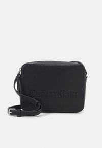 Borsa a tracolla in pelle nera con forma rettangolare, chiusura con zip, tracolla regolabile e marchio "Calvin Klein" traforato sul davanti.
