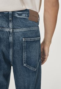 Jeans in denim blu scuro con una toppa del logo marrone sulla fascia posteriore in vita, con tasche standard e un design a gamba dritta.