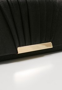 Pochette noire avec un design plissé, fabriquée en tissu texturé. Dispose d'un fermoir rectangulaire en ton doré pour la fermeture.