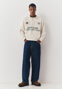 Jonge man met handen in zakken, gekleed in een crèmekleurige, getextureerde trui met zwarte tekst en losse donkerblauwe jeans, staand tegen een witte muur.