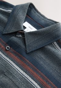 Camicia blu navy con trama e righe fantasia in rosso, bianco e grigio. Presenta una chiusura con bottoni e un colletto appuntito. Etichetta del marchio visibile.