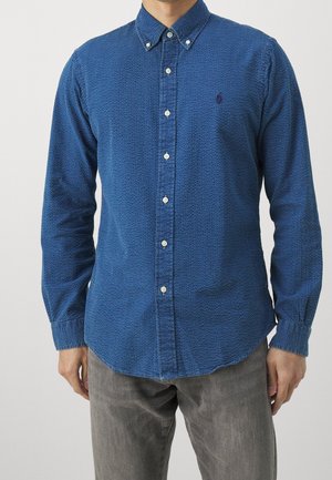 Blue patterned button-up shirt with a collar, long sleeves, and subtle texture; features white buttons and a small logo on the chest.