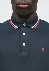 Ναυτικό πουκάμισο πόλο με υφαντό ύφασμα, διαθέτει τρία κουμπιά στην μπροστινή πλευρά, γιακά με κόκκινες λεπτομέρειες και ένα μικρό κόκκινο λογότυπο στο στήθος.