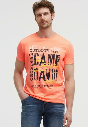 Mann trägt ein knallorangfarbenes T-Shirt mit kurzen Ärmeln und schwarzem sowie gelbem Schriftzug "Outdoor Abenteuer", kombiniert mit blauen Jeans, vor einfarbigem Hintergrund stehend.