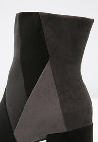 Wildleder-Ankle-Boot in Schwarz und Grau, mit geometrischer Panele und einer glatten Textur. Das Design betont moderne Linien und klare Formen.