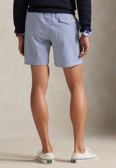 Osoba ubrana w niebiesko-białe prążkowane szorty, granatowy sweter i białe trampki, stojąca tyłem do kamery na neutralnym tle.