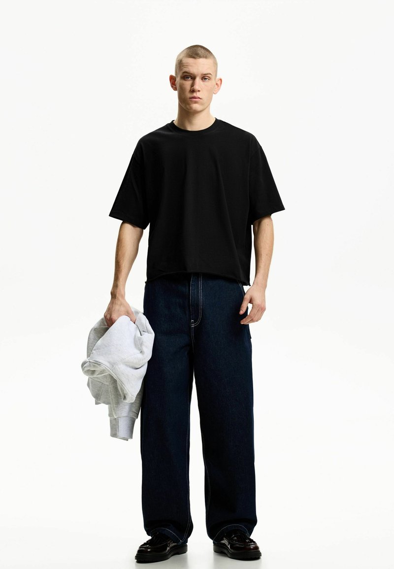 Tricou negru cu mânecă scurtă asociat cu blugi dark denim cu pantaloni largi. În mână este un hanorac gri deschis. Design minimalist, stil casual.