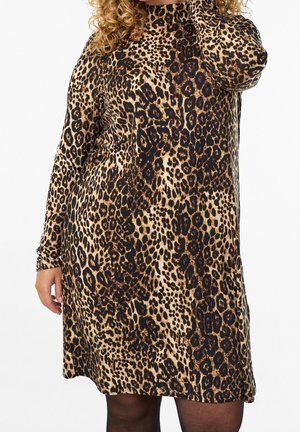 Personne aux cheveux bouclés portant une robe à manches longues, longueur genou, à imprimé léopard avec des collants noirs sur fond blanc.