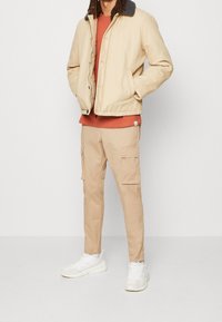 Beżowa kurtka ocieplana z ciemnym kołnierzem, T-shirt w kolorze rdzy, beżowe spodnie cargo i białe sneakersy, stojąca na tle jednolitego tła.