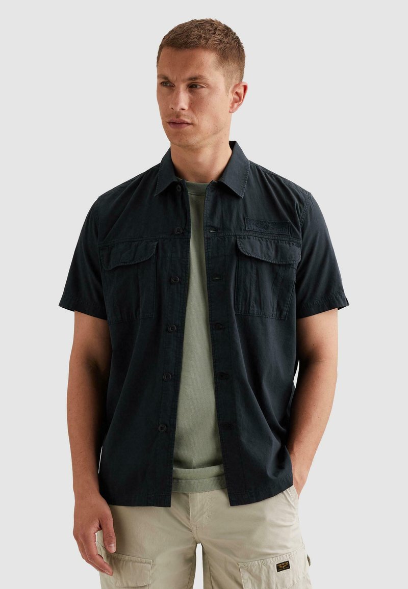 Homme portant une chemise noire à manches courtes avec boutons par-dessus un t-shirt vert, un short cargo beige, debout avec une main dans la poche, regardant vers la gauche.