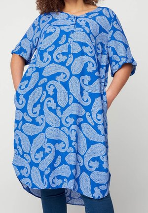 Donna che indossa una tunica blu larga con motivo paisley azzurro chiaro e tasche, abbinata a jeans blu scuro.
