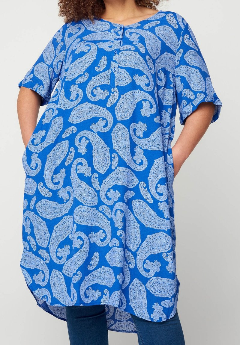 Femme portant une tunique bleue ample avec un motif cachemire bleu clair et des poches, associée à un jean bleu foncé.