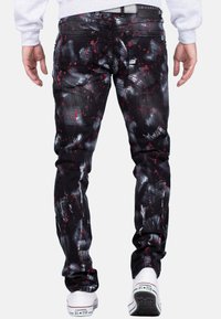 Jeans in denim nero con motivi di spruzzi rossi e bianchi, design aderente, cinque tasche e sneaker bianche in vista.