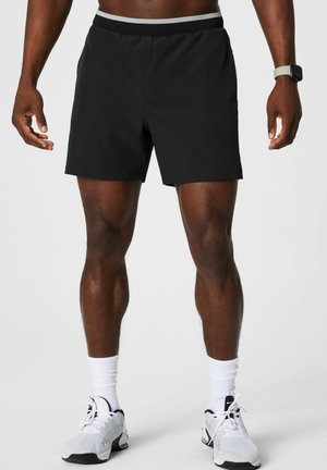 Shorts de sport noirs avec une taille élastique, présentant un design minimaliste et une texture lisse, portés avec des chaussettes de sport blanches et des chaussures.