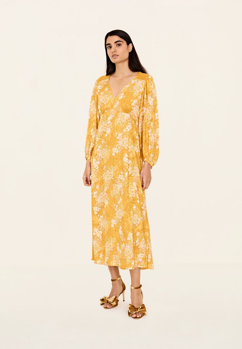 Rochie lungă galbenă cu flori, de tip V la decolteu, cu mâneci lungi bufante și un design fluid. Complementată de sandale cu toc înalt, maro, cu bretele.