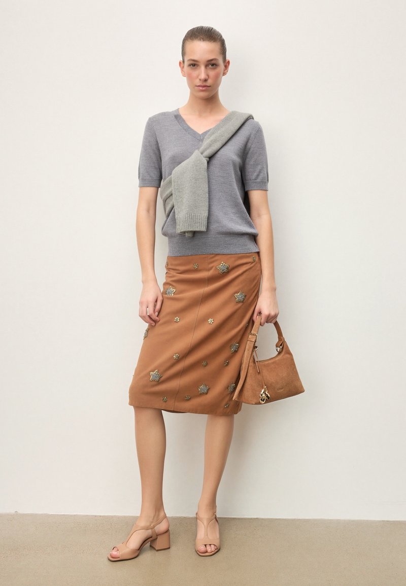 Femme portant un pull gris à manches courtes avec une écharpe assortie, une jupe marron ornée, des sandales nude à talons blocs, et tenant un sac à main en daim beige.