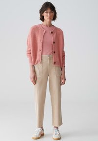 Hellrosane Strickjacke aus strukturierter Stoffqualität mit Knöpfen, über einem pinken Hemd getragen. Kombiniert mit beigen Hosen und weißen Sneakers.