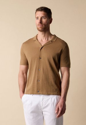 Mann trägt kurzärmliges braunes Hemd mit Knöpfen und weiße Hose, steht mit Händen in den Taschen, neutraler beiger Hintergrund.