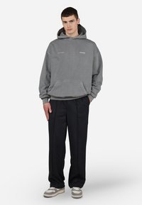 Grå hættebluse med kangaroo-lomme, sammen med sorte bukser med et diskret logo, og lysegrå sneakers med hvide detaljer.