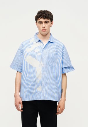 Noor mees, kes kannab lahtist sinise ja valge vertikaalse triibuga lühikest varrukat särki ees oleva rinnatasku ja mustade pükstega.