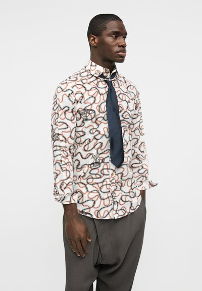 Άνδρας που φοράει λευκό πουκάμισο με μοτίβο αφηρημένων μαύρων και πορτοκαλί γραμμών, γραβάτα σε ναυτικό μπλε και φαρδιά σκούρα γκρι παντελόνια.