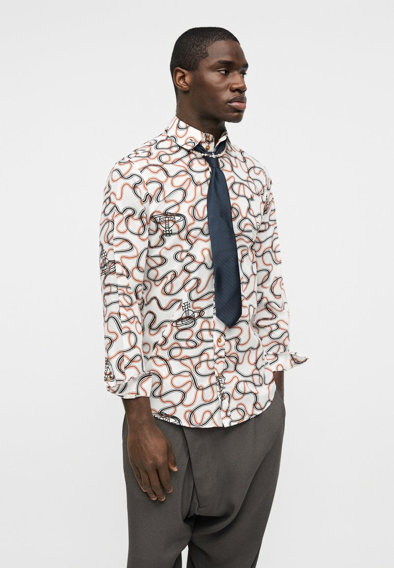 Uomo che indossa una camicia bianca con bottoni e un motivo astratto a linee ondulate nere e arancioni, cravatta blu navy e pantaloni larghi grigio scuro.