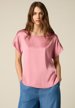 Donna con capelli mossi che indossa una camicetta rosa lucida a maniche corte e pantaloni blu, in piedi con le mani in tasca su uno sfondo semplice.