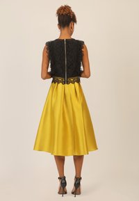 Schwarzes Spitzenoberteil mit gewelltem Saum, gelber Satinrock, Reißverschluss hinten und schwarzen Absätzen. Glatte Textur und figurbetonter Taillenschnitt.